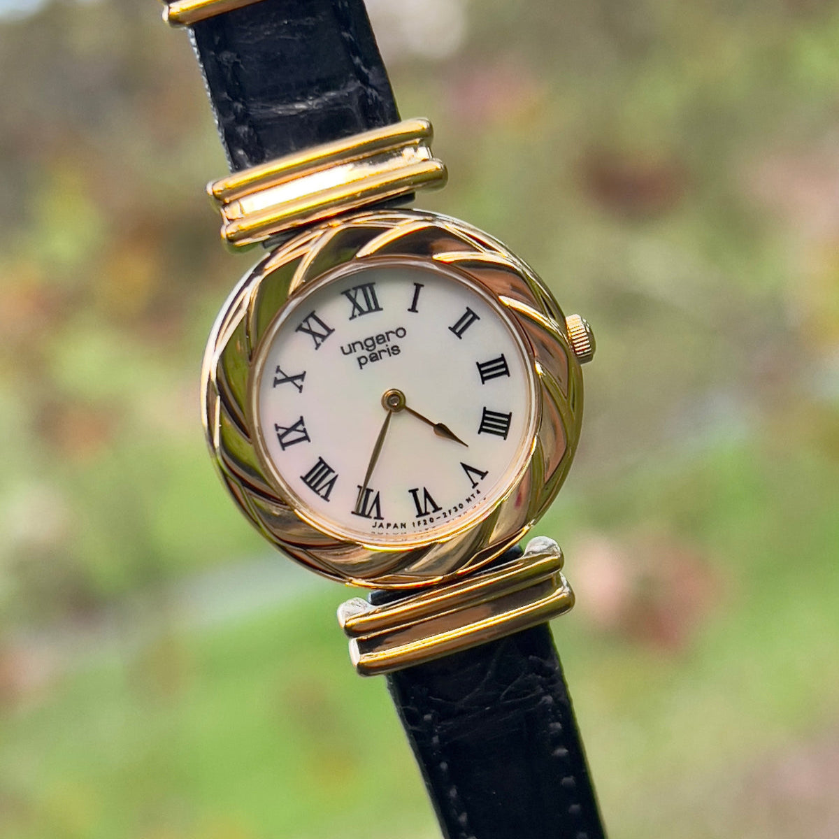 A Ungaro Paris woman's watch with a pearl dial, gold-tone case, and black leather strap with decorative buckles.