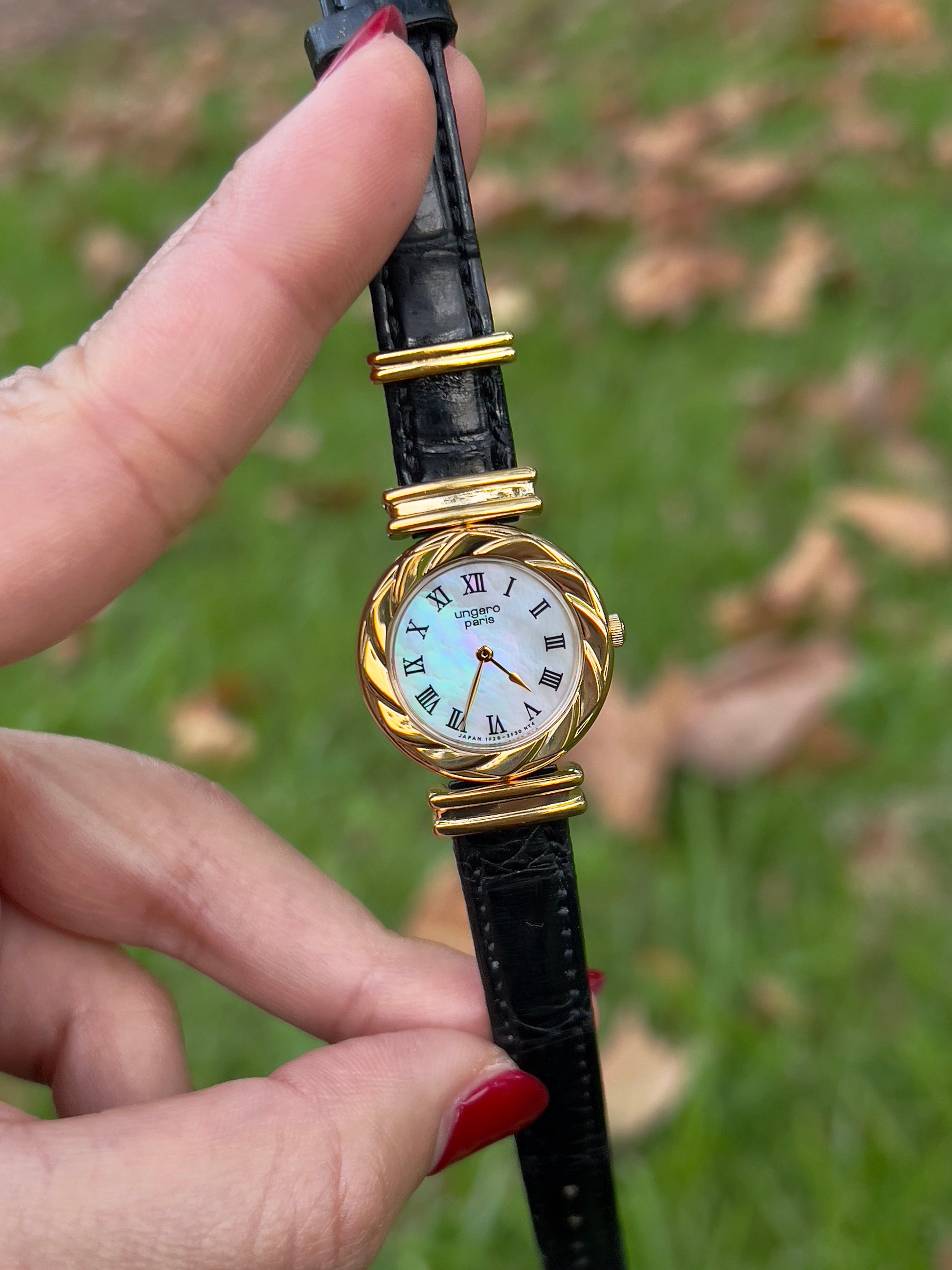 Ungaro gold and black wristwatch held by a hand with red nail polish against a blurred natural background.