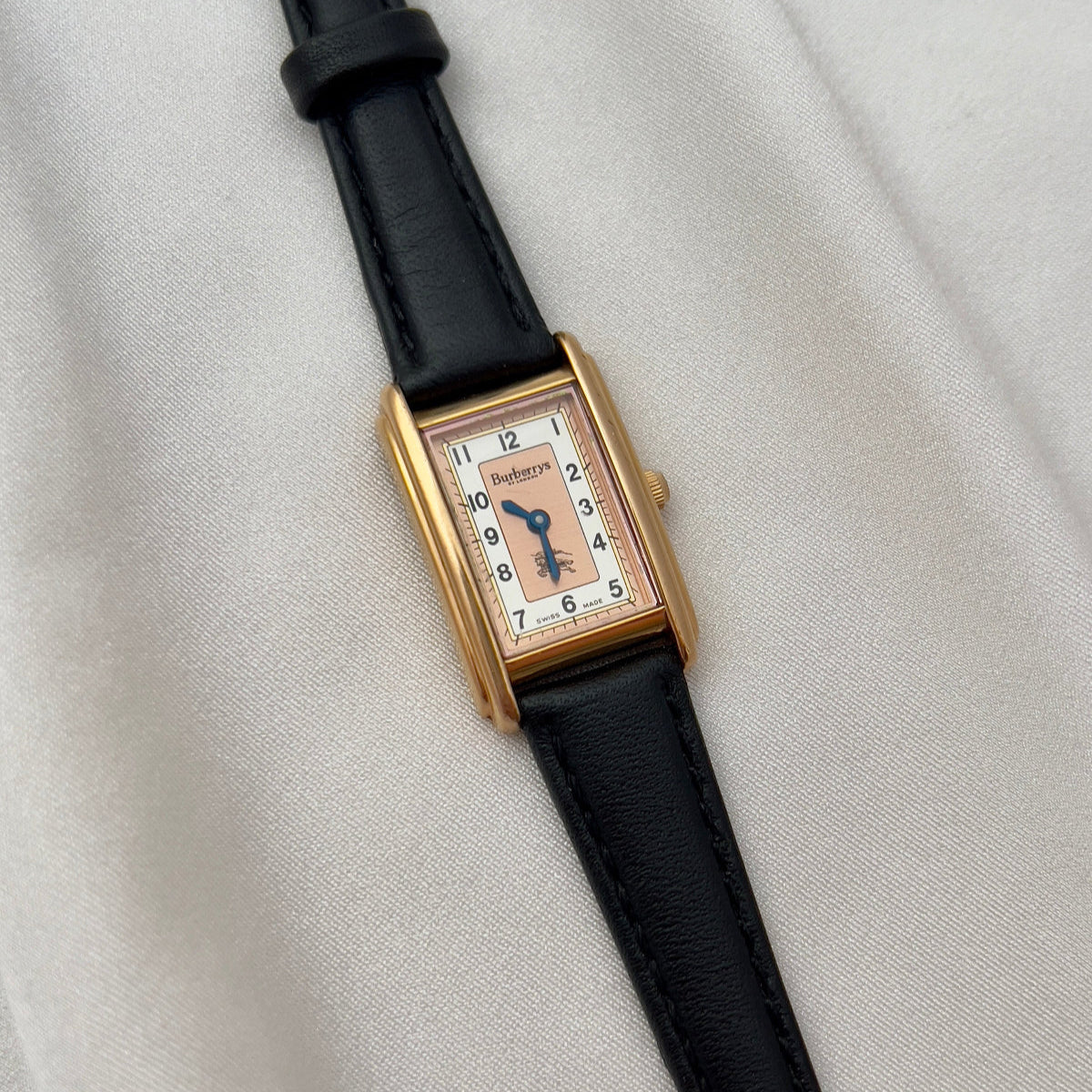 Rectangular watch with gold case and black strap on a light gray background