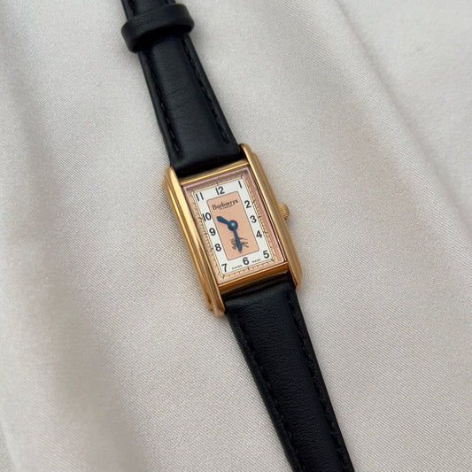 Rectangular watch with gold case and black strap on a light gray background