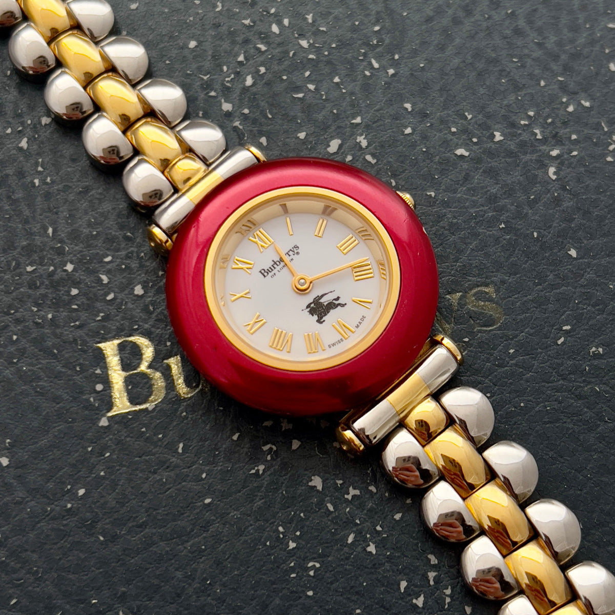 Burberry's of London woman's watch with red face and gold and silver band on a black textured surface
