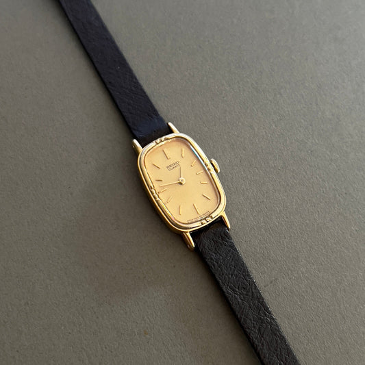 Gold Seiko Quartz luxurious woman's watch with black strap on a gray background