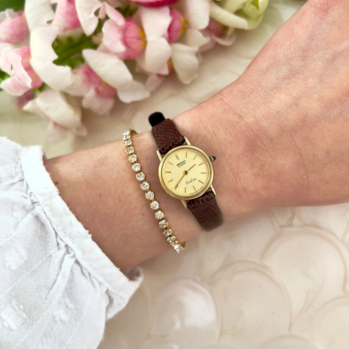 Seiko Exceline gold ladies' watch and brown leather bracelet on a wrist with flowers in the background