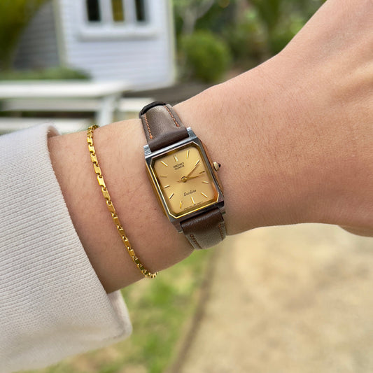 Vintage Seiko Exceline ladies gold tank watch with double-framed rectangular case and brown leather strap, styled on herringbone tweed fabric.