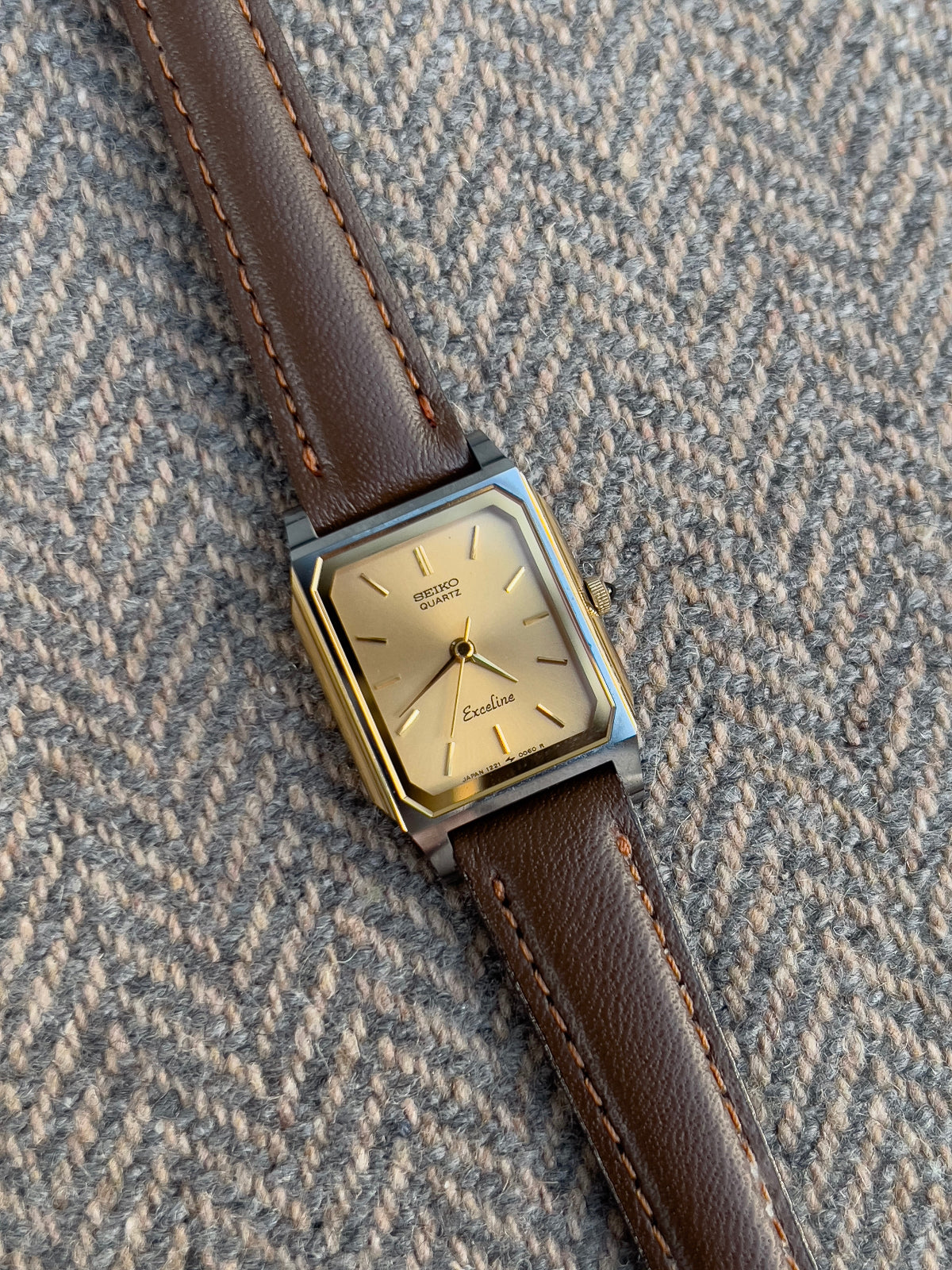Vintage Seiko Exceline ladies gold tank watch with double-framed rectangular case and brown leather strap, styled on herringbone tweed fabric.