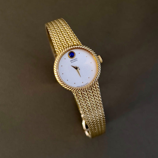 Vintage Seiko 1220-0020 ladies gold tone watch with cream dial, blue cabochon at 12 o’clock, and integrated bracelet