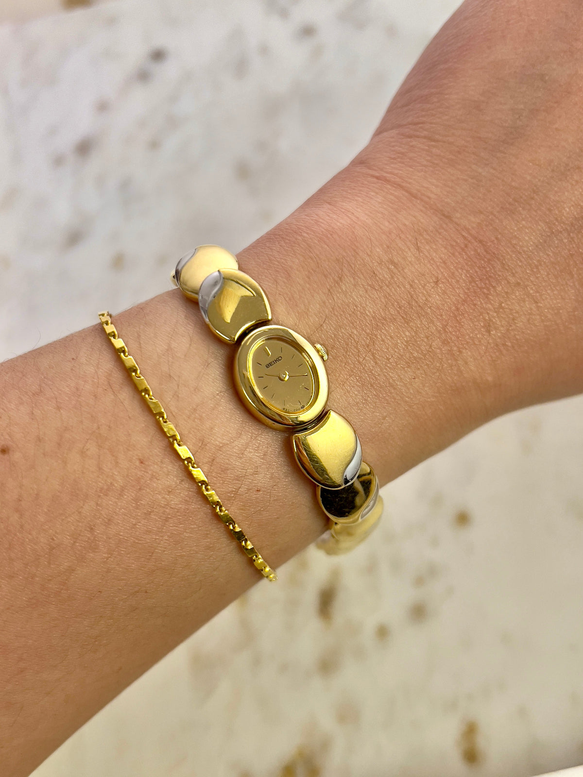Gold bracelet with a watch face on a wrist against a neutral background
