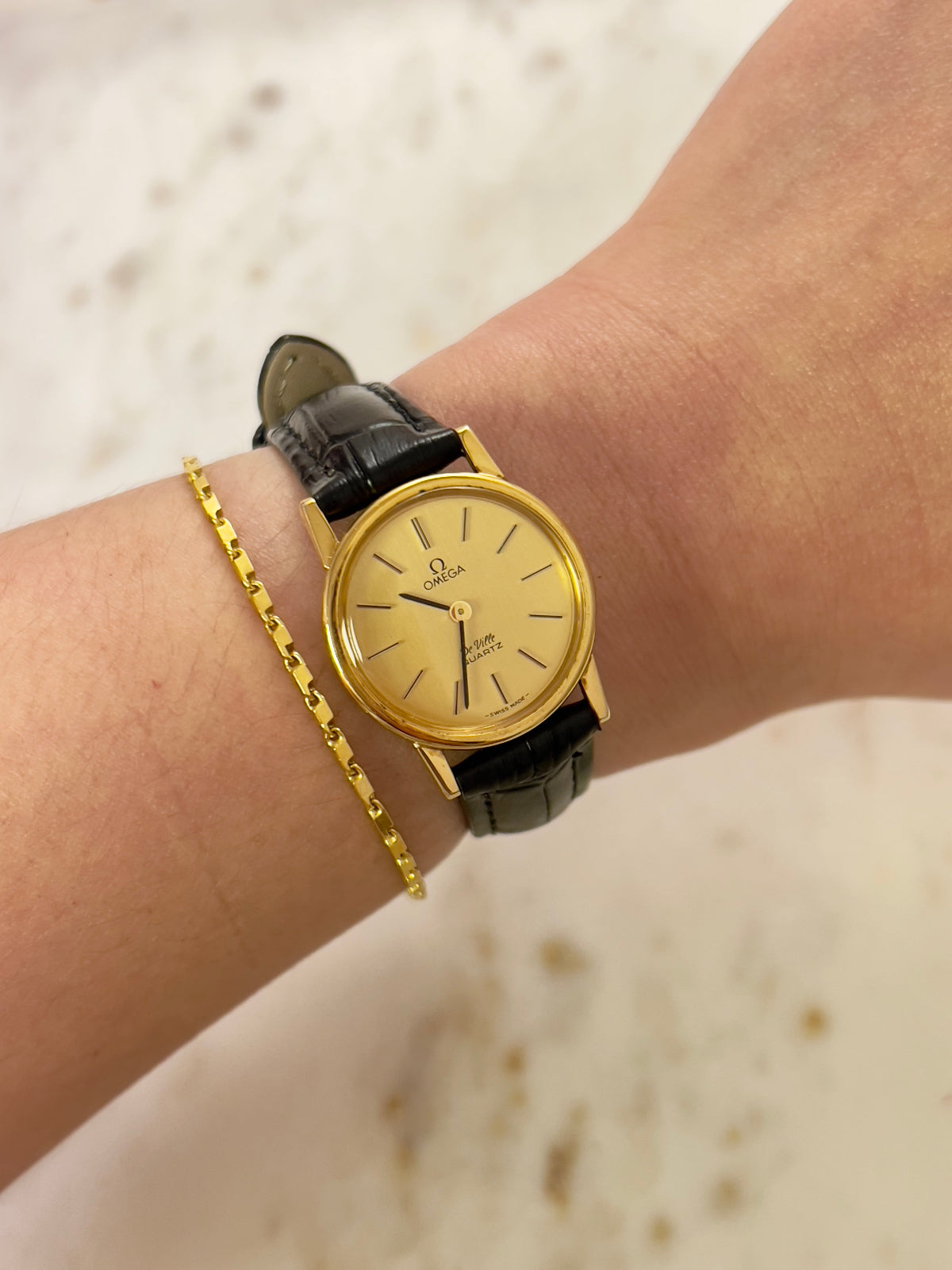 Omega gold watch with black strap on a wrist against a neutral background