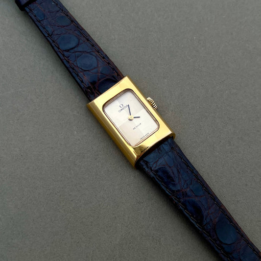 Omega Woman's Wristwatch with gold case and blue leather strap on a gray background De Ville Vintage Watch NZ