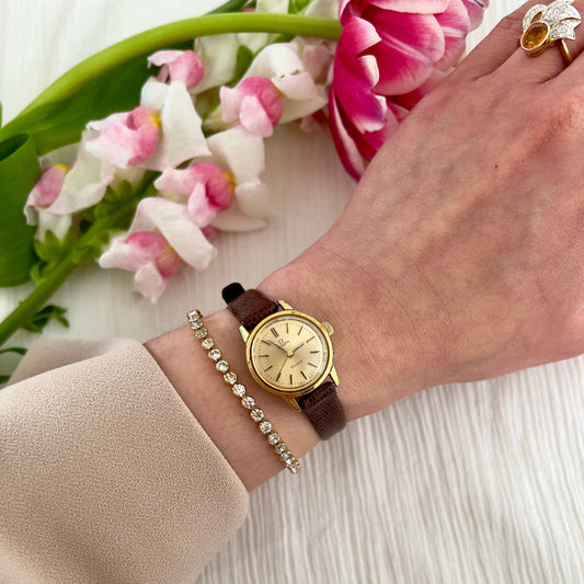 Vintage Omega ladies gold watch on wrist with brown leather strap and champagne dial, styled with diamond bracelet and flowers – elegant Swiss dress watch for women in New Zealand.