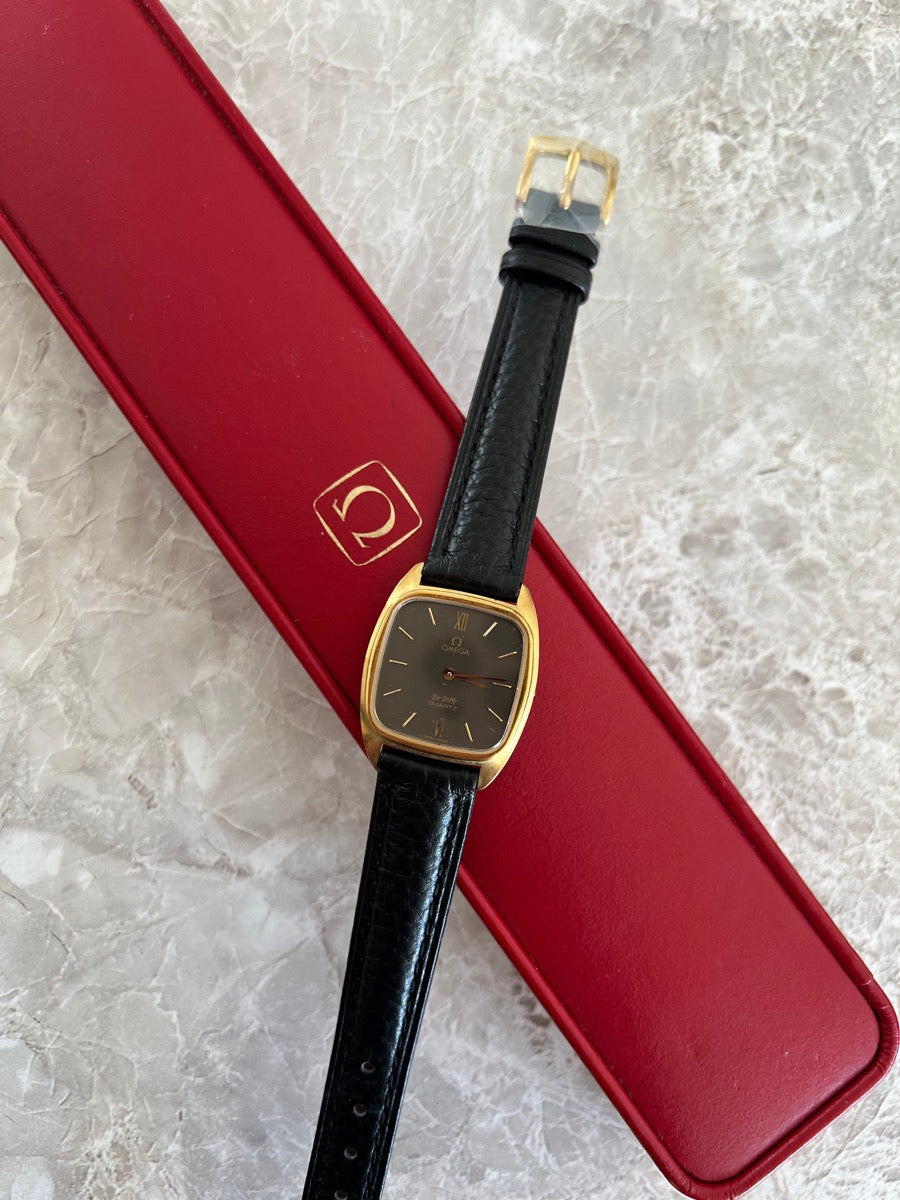 Omega De Ville 591.0099 with dark dial and black leather strap beside a red Omega-branded case