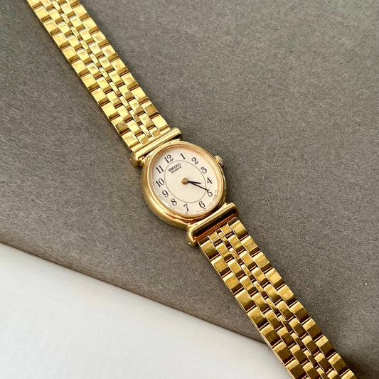 Seiko ladies gold bracelet watch with ivory dial and black numerals on wrist beside diamond tennis bracelet
