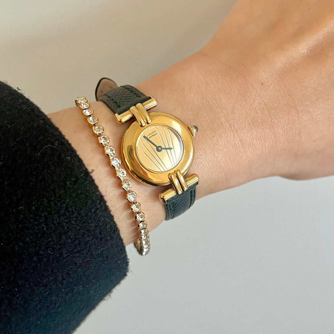 Gold Cartier Colisée sunray dial watch on a dark leather strap worn beside a fine bracelet