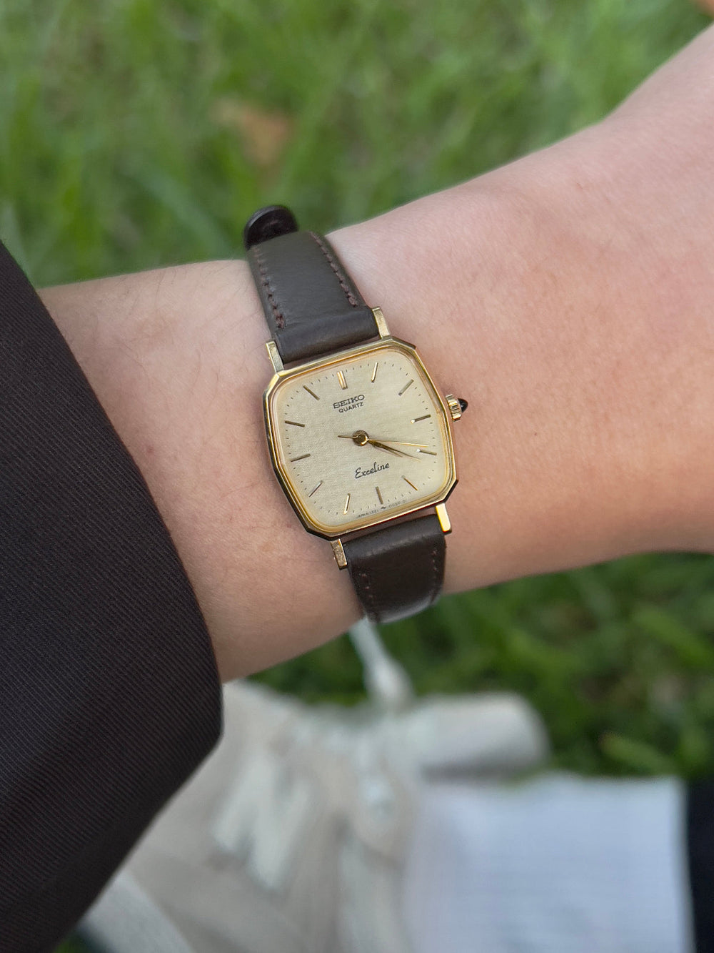 Vintage Seiko Exceline gold-tone watch on a brown leather strap worn below a clean black cuff