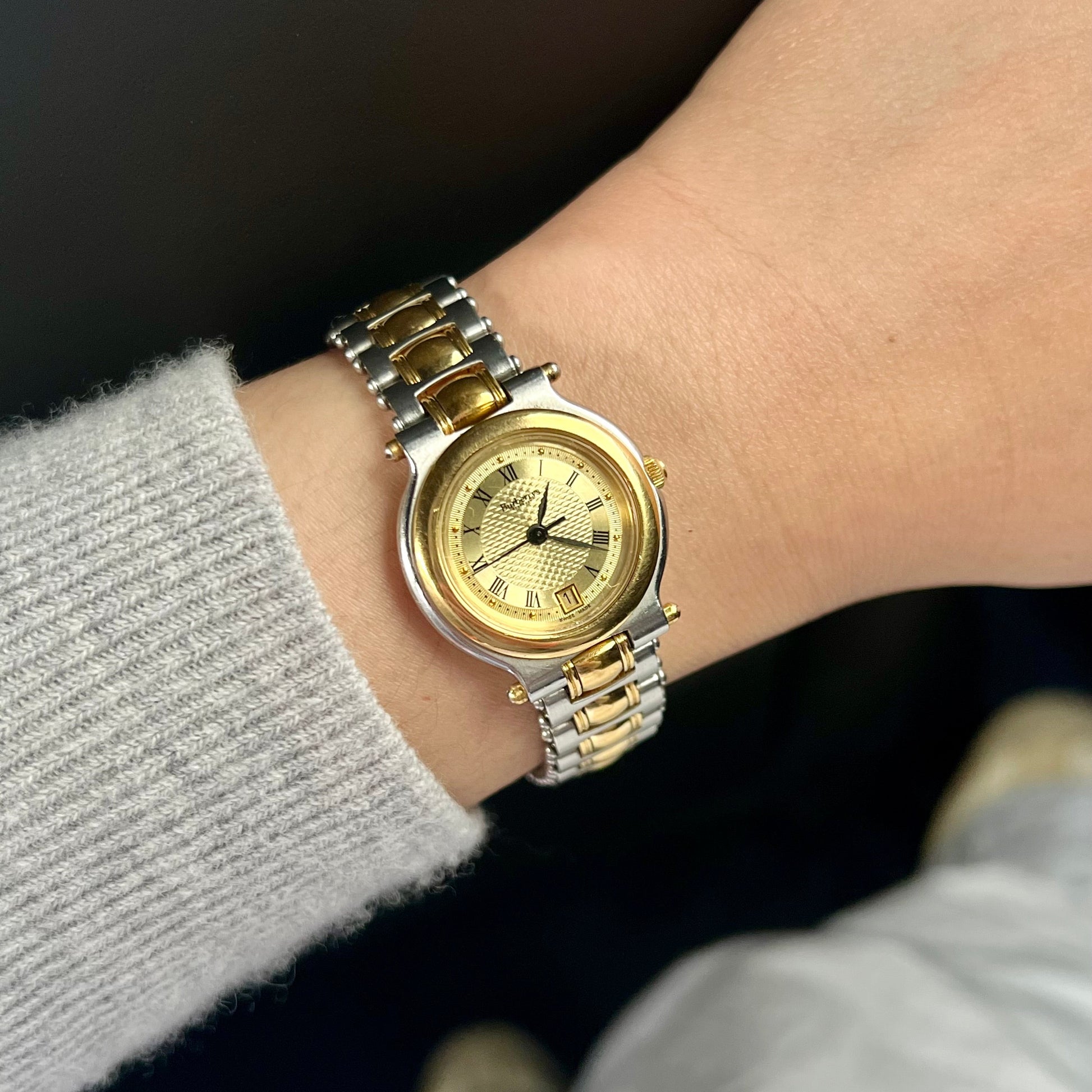 A two-tone Burberry Life gold and silver metal wristwatch with a gold dial, worn on a person's wrist with a light gray knit sweater sleeve visible.