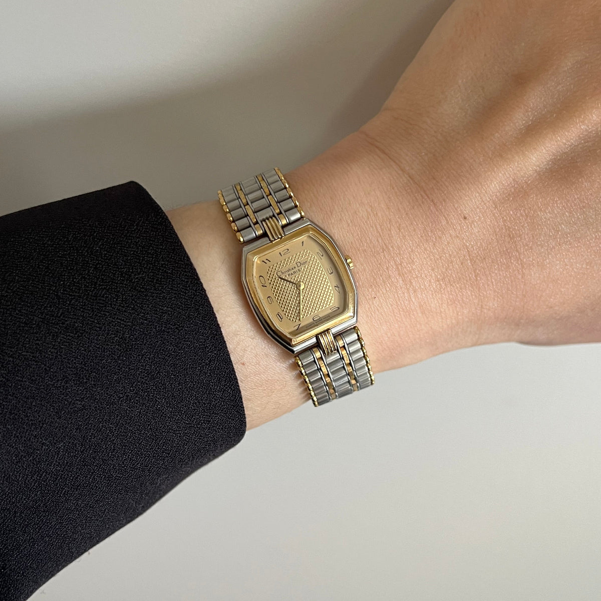 A two-tone metal wristwatch with a gold-toned dial and matching gold-toned bezel, worn on a person's wrist with a black sleeve visible.