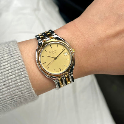 A wrist wearing a Christian Dior two-tone metal watch with a gold dial and date window, photographed against a white garment and dark background.