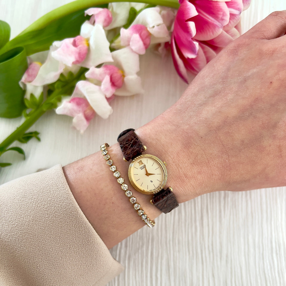 Wrist wearing a watch with a bracelet, surrounded by pink and white flowers on a light background