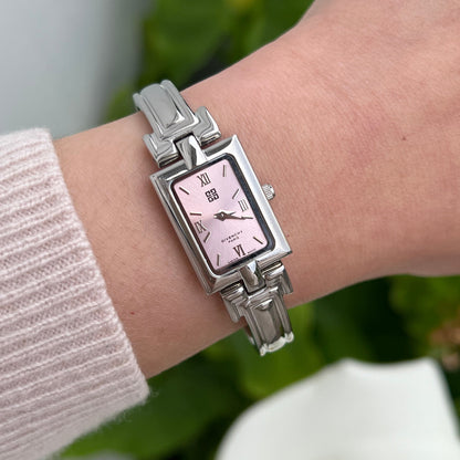 A silver-toned rectangular metal watch with a pink dial and Roman numeral hour markers, shown worn on a person's wrist. The watch has a matching silver link bracelet and is photographed against a soft-focus background of green leaves and white flowers.