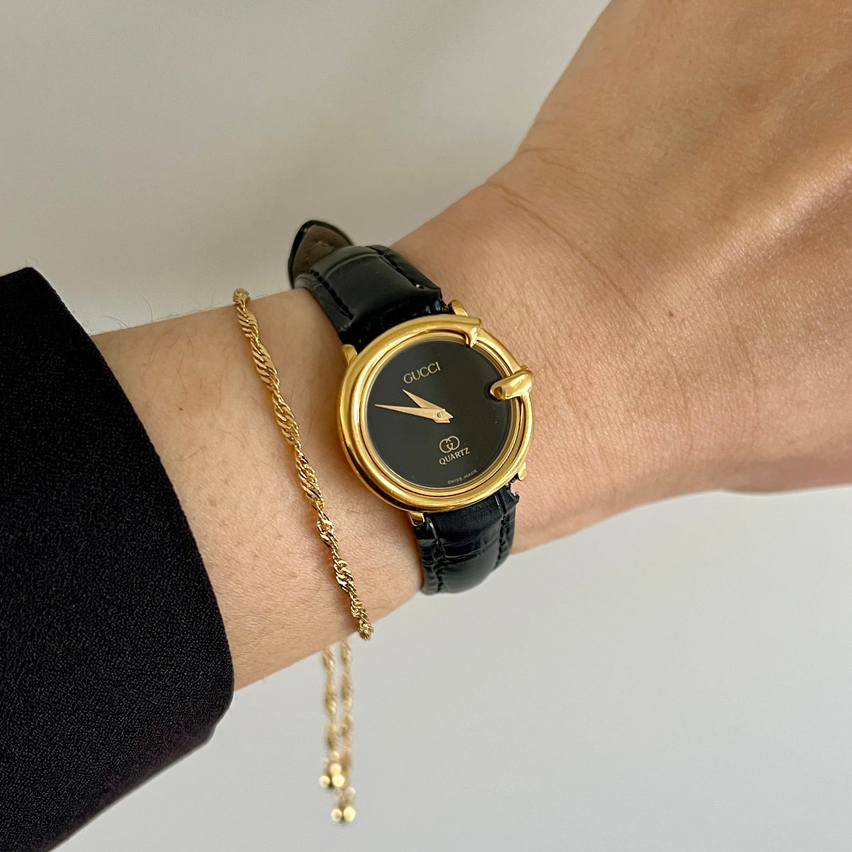 A close-up of a person's wrist wearing a Gucci watch with a gold bezel, black dial, and black leather strap, alongside a gold chain bracelet, against a plain light background.