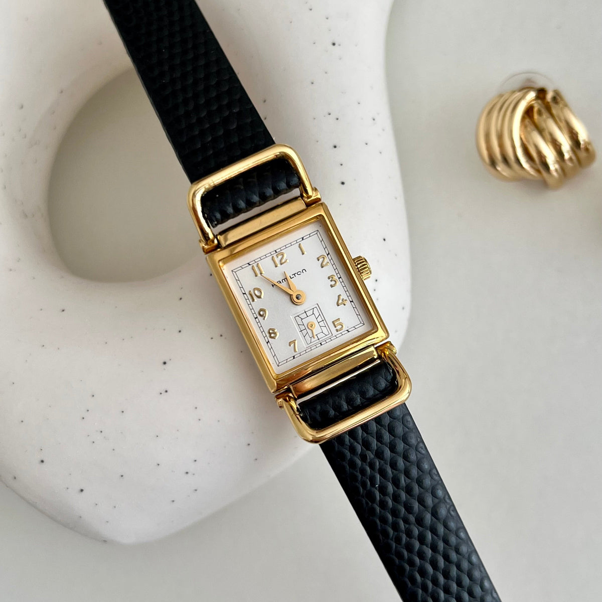 Hamilton Tank Women's watch with gold case and black strap on a textured white surface