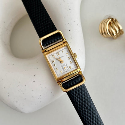 Hamilton Tank Women's watch with gold case and black strap on a textured white surface