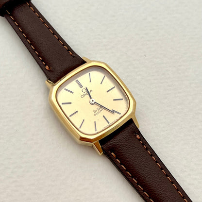 Omega De Ville Cushion Case with gold case and brown leather strap on a light gray background