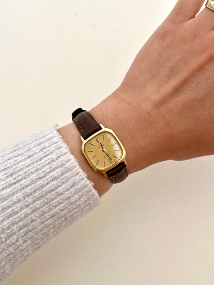 Gold watch with brown leather strap on a wrist against a beige background