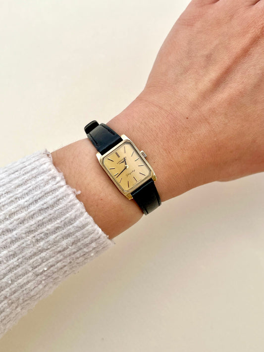 Hand wearing a gold rectangular Longines Women's watch with a black strap on a light background