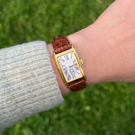 Gold Hamilton watch with a brown leather strap on a wrist against a grassy background
