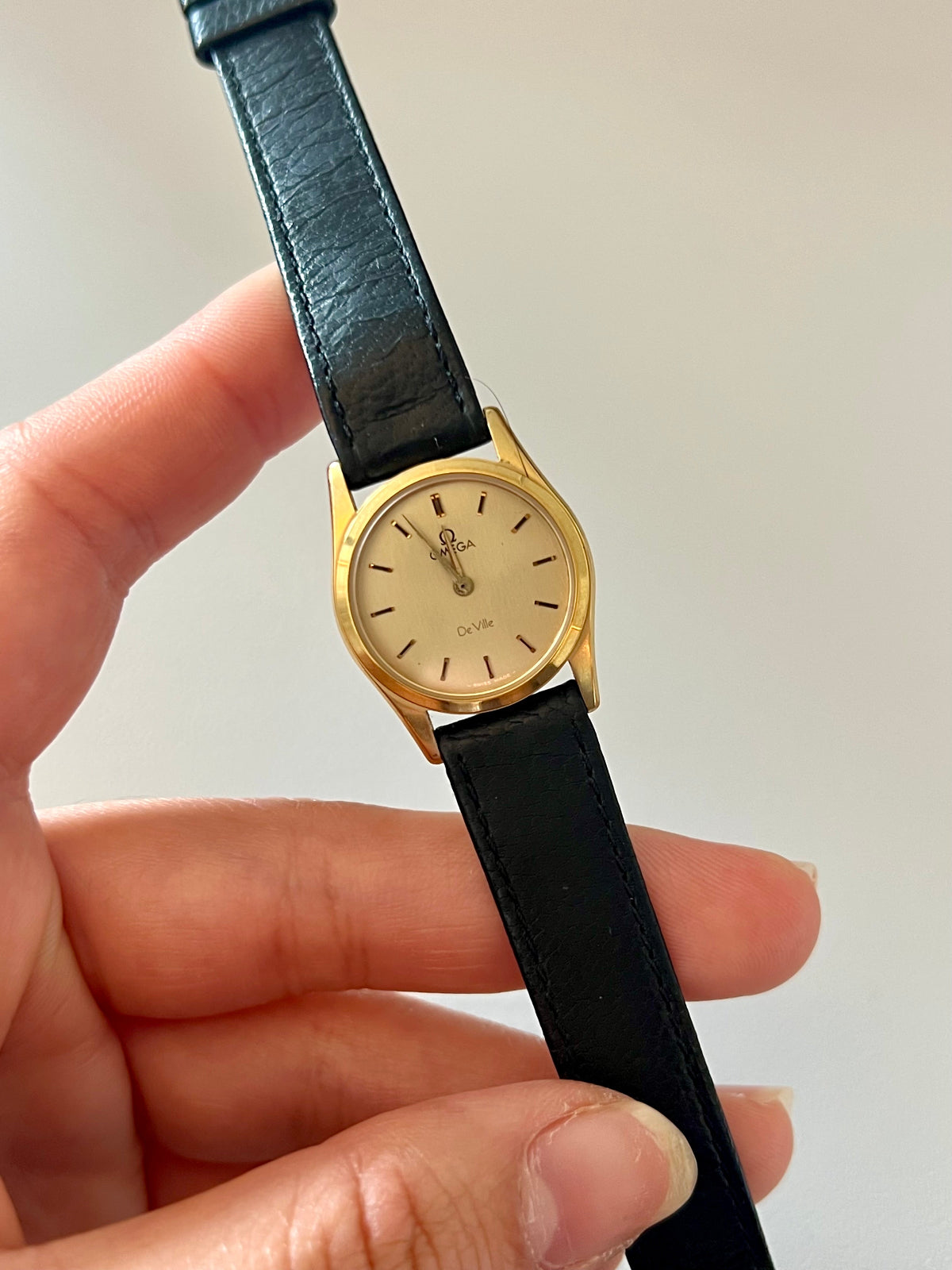 Gold Omega Women's watch with black leather strap held by a hand against a plain background