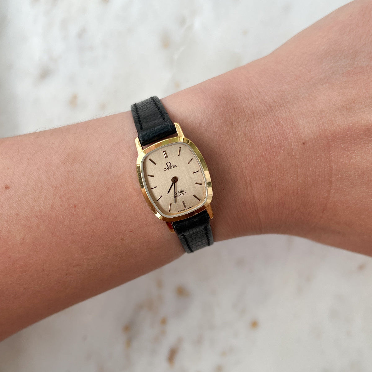 Gold watch with black strap on a wrist against a light background