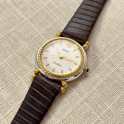 A vintage Seiko Neoclassical styled two-toned woman's watch with a gold-tone bezel and a brown leather strap, displaying a champagne dial and gold-accented lugs.