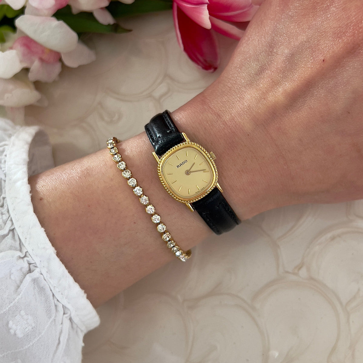Gold Rado ladies watch with black strap on wrist, bracelet, and flowers in the background