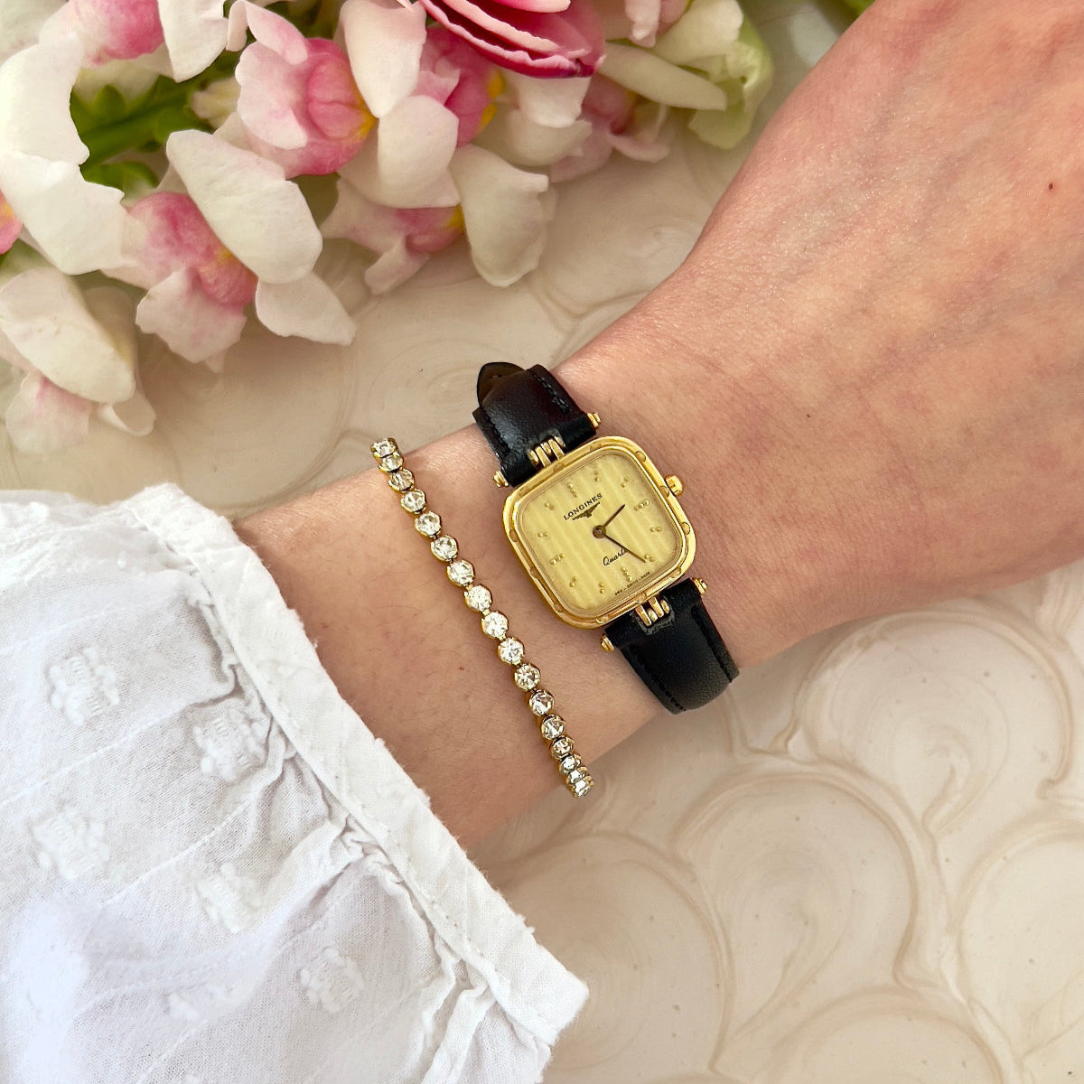 Gold Longines ladies' watch on a wrist with a pearl bracelet, against a floral background