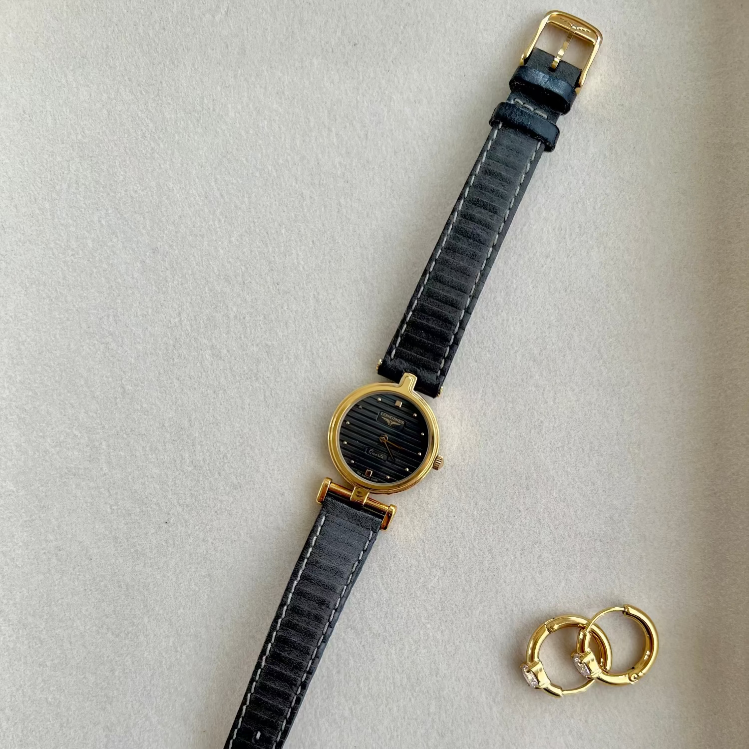 Black Longines Round Women's watch with gold accents and a small gold ring on a light gray background
