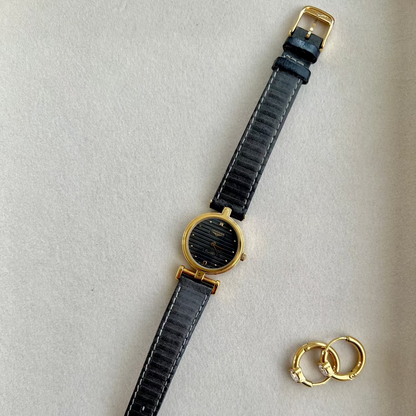 Black Longines Round Women's watch with gold accents and a small gold ring on a light gray background