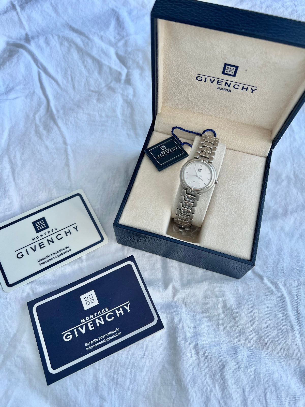 A Givenchy watch with a silver-toned metal bracelet and a white dial, presented in an open navy blue and beige Givenchy box on a white fabric background, alongside its branded card and box lid.