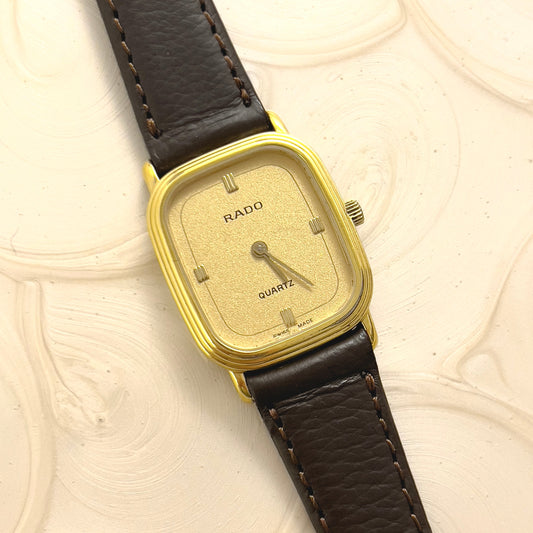 Rado vintage woman's watch with a gold case and black leather strap on a textured beige background