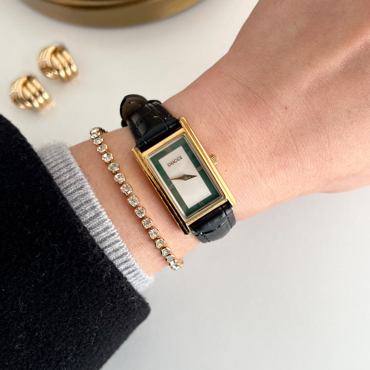 A close-up lifestyle shot of a Gucci watch on a person's wrist, styled with a gold bracelet and earrings on a clean white surface.