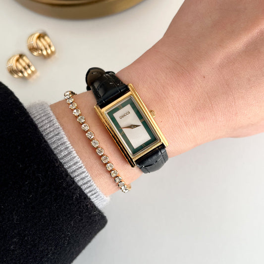 A close-up lifestyle shot of a Gucci watch on a person's wrist, styled with a gold bracelet and earrings on a clean white surface.