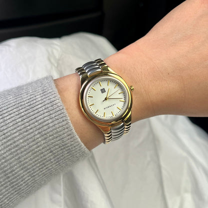 A two-tone Givenchy stainless steel and gold analog watch with a white dial, shown worn on a person's wrist over a gray ribbed sweater sleeve and white fabric background.