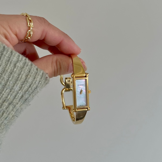 A gold-tone Gucci 1500L wristwatch with a rectangular face and white dial, held by a hand wearing a gray knit sweater and a gold chain ring against a plain light gray background.