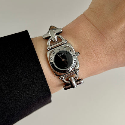 A silver-tone Gucci bracelet watch with a black dial and rhinestone hour markers, shown worn on a wrist with a black sleeve visible.