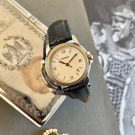 Tissot watch with gold bezel and black leather strap on a textured surface with a book and gold hoop earrings.