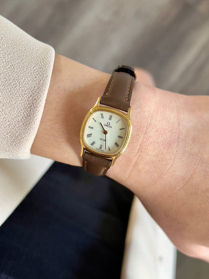 Omega Gold luxurious woman's watch with a brown leather strap on a wrist against a neutral background