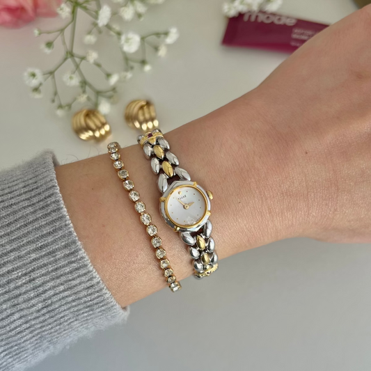 A two-tone gold and silver Seiko Tisse watch with a white dial is worn on a wrist alongside a matching gold bracelet and a crystal-studded bracelet, with baby's breath flowers and a small purple packet in the background.