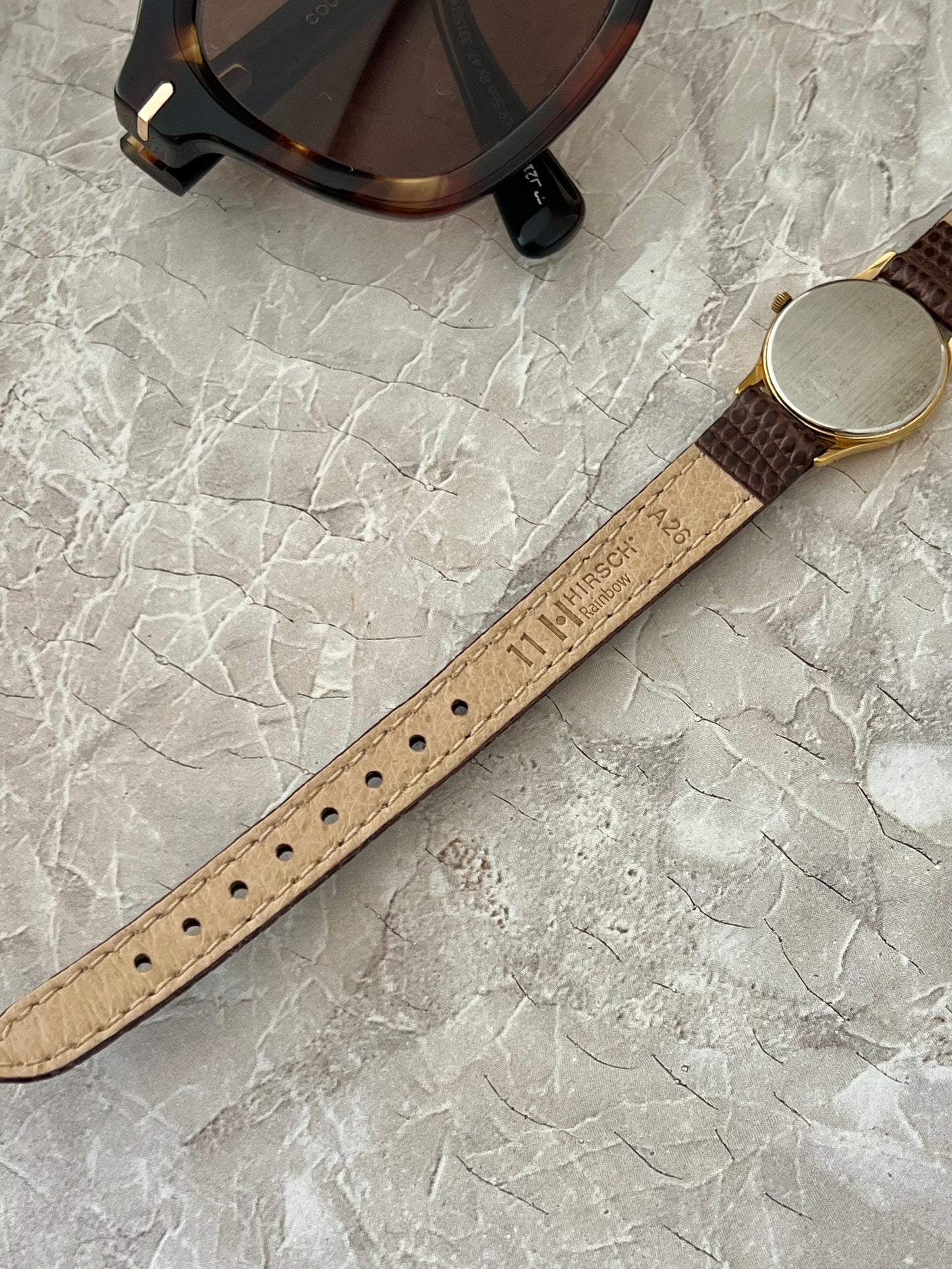 A close-up of a vintage-style wristwatch strap with a tan leather inlay and dark brown Hirsch crocodile-embossed ends, laid diagonally on a light gray marble surface next to a pair of sunglasses.