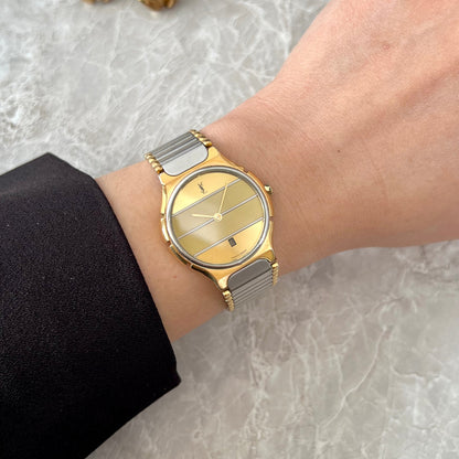 A gold and silver two-tone watch with a striped dial, worn on a person's wrist. The watch has a gold bezel, gold-toned striped dial, and a silver-toned link bracelet.