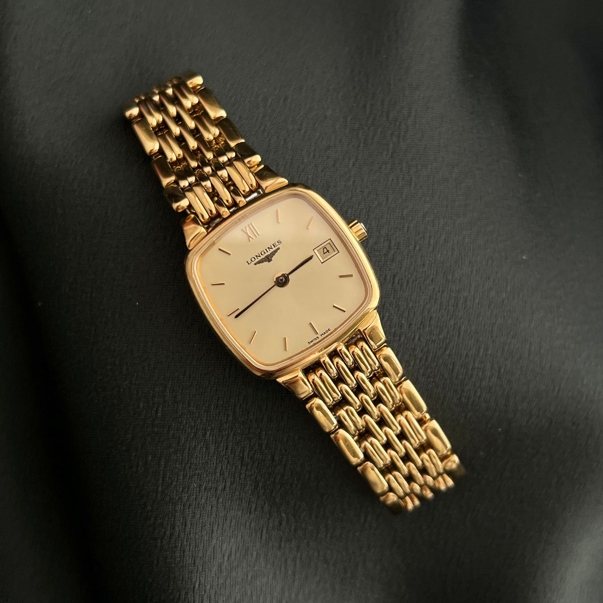 A gold-tone wristwatch with a square dial and matching metal link bracelet, photographed on a dark textured fabric background.